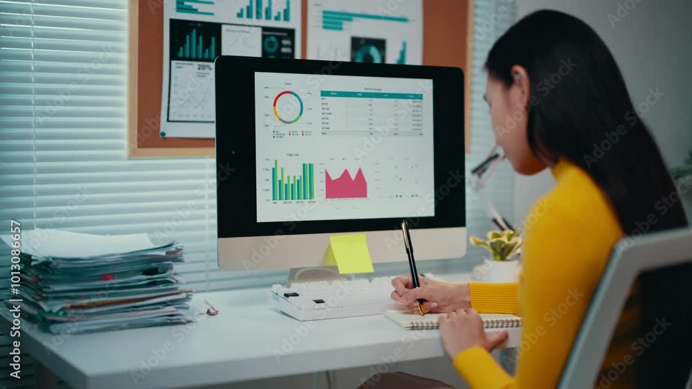 Poster young asian businesswoman analyzes financial data on a computer in her office, taking notes in a not