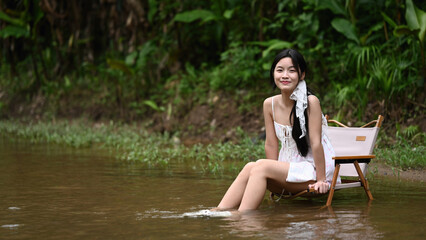 A teenage girl sits on a small chair, dipping her feet in a calm stream, smiling and enjoying a peaceful moment in nature