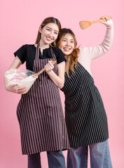 Photo two cute happy funny young Asian beautiful female, woman smiling wearing aprons holding baking equipment makes homemade cake poses isolated plain pastel light over pink color wall background.