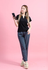 Fototapeta premium Full length photo cute happy fun young Asian beautiful female, woman smiling in a black t-shirt and holding a mobile phone enjoy poses isolated plain pastel light over pink color wall background.