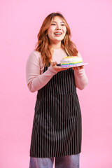 Photo cute happy fun young Asian beautiful female, woman smiling wearing aprons holding cake sweet dessert licking fingers poses isolated plain pastel light over pink color wall background.