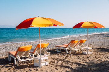 Bright relaxing travel background. Two lounge chairs and red and yellow beach umbrella on sandy sea coast in early morning. Copy space for your text. Holiday vacation on the seaside.