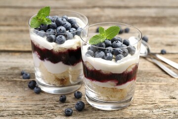 Tasty trifle dessert. Sponge cake, blueberries, jam and whipped cream in glasses on wooden table, closeup
