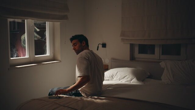 Guy browsing cellphone late evening in dark bedroom. Sleepless man on night bed