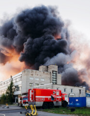 Fototapeta premium Massive large blaze fire in the city with large column plume of smoke, blazing warehouse factory, storage building is burning, putting out the fire, extinguish the fire, arson, burning house damage