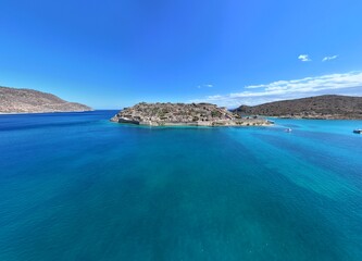 This aerial drone photo captures the beautiful Plaka Marina with its charming fishing boats, set against the backdrop of Spinalonga Island off the coast of Crete, Greece. The image highlights the