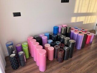 Colorful foam massage rollers neatly arranged in rows on a wooden floor against a white wall, ready for fitness or physiotherapy use.
