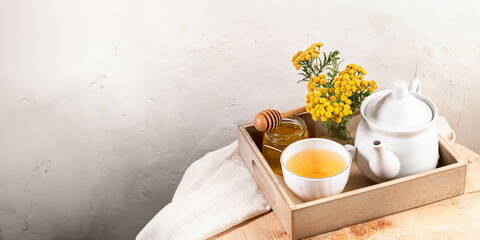 Tansy tea in teapot and cup, tansy flowers, honey in wooden tray on textured white wall backdrop.