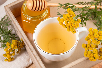 White cup with healthy medicine tansy tea, honey and tansy flowers in wooden tray close up.