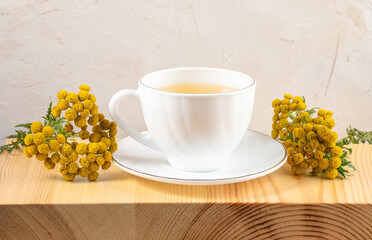White cup on saucer of healthy herbal tansy tea and tansy flowers on textured wooden board close up.