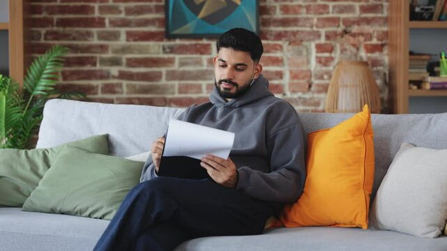 Serious man wearing casual clothes and leafing through papers on black tablet with concentrated expression. Young male filling out paperwork and placing folder aside on grey sofa with bright pillows.