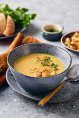 Bowl of mashed potato soup with a spoon on the plate. The bowl is filled with yellow soup puree and topped with croutons, parsley and ground red pepper.