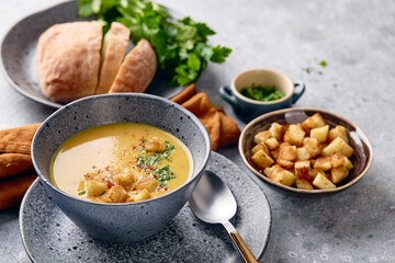 Bowl of mashed potato soup with a spoon on the plate. The bowl is filled with yellow soup puree and topped with croutons, parsley and ground red pepper.
