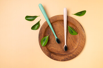 Multicolored eco-friendly bamboo toothbrushes with green leaves on wooden plate on beige background. Natural accessories for oral hygiene, no plastic. Dental health concept top view flat lay