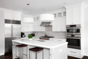 Fototapeta premium A white kitchen with pendant lights hanging above a large island, stainless steel appliances, and a grey subway tile backsplash. No brands or labels.
