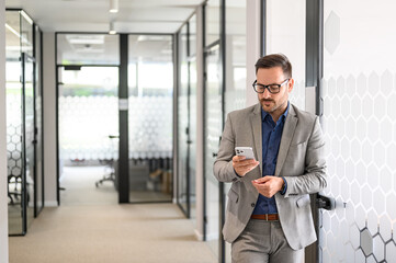 Serious young man in eyeglasses messaging online over mobile phone while standing in office corridor