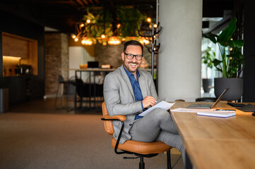 Positive young businessman in eyeglasses smiling at camera and working over laptop at desk in office