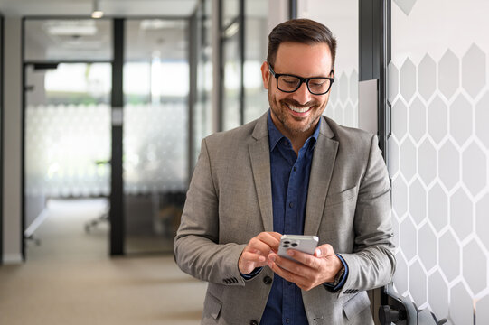 Successful male entrepreneur smiling and checking messages over mobile phone in office corridor