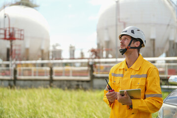young caucasian technician in hardhat holding laptop computer and walkie talkie while inspection in oil refinery industry