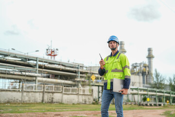 young caucasian technician in hardhat holding laptop computer and walkie talkie while inspection in oil refinery industry