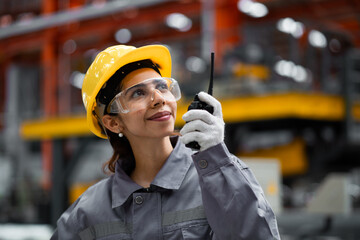 A woman wearing a yellow helmet and safety glasses is holding a walkie talkie. She is smiling and she is happy