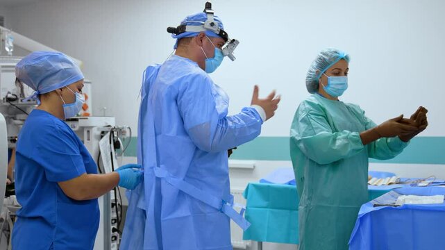 Caucasian surgeon waves his hands to dry them up after washing and sanitizing. Nurses tie doctor's robe and prepare latex gloves.