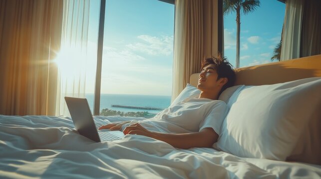 A man in a white shirt lies in bed and looks out the window at the ocean as the sun rises. He has a laptop on his lap. - Powered by Adobe