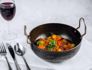 Spicy chicken curry and vegetable dish featuring vibrant bell peppers, and eggplant. Garnished with fresh herbs. Sits on a clean white tablecloth. A glass of red wine sparkles next to the dish.