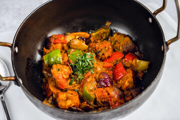 Spicy vegetable and chicken curry dish in a traditional metal cooking pot. It includes bright red and green bell peppers, chunks of tomatoes, and herbs, all glistening with spices.