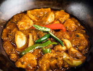 Spicy chicken curry served in a dark stone bowl, with chicken pieces in a vibrant, aromatic sauce, garnished with green and red chilies and onion chunks.