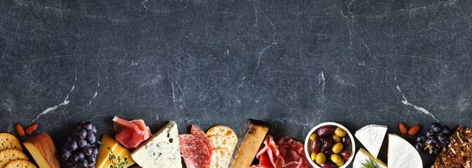 Variety of charcuterie cheeses, meats and appetizers. Top view bottom border on a dark black stone...
