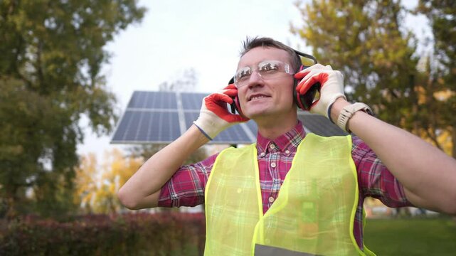 Man wearing hearing protection safety equipment at solar power station. Engineer wears noise canceling headphones or earphones against of solar panel. Foreman puts on protective ears with headphones.