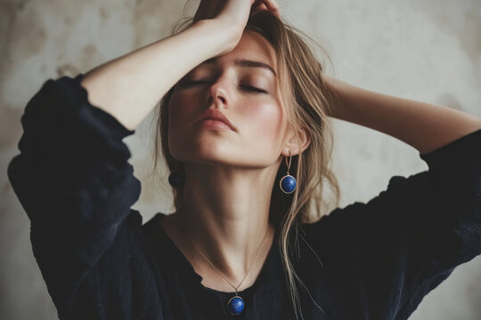 Blonde woman wearing blue earrings and a necklace with lapis lazuli stone. Her eyes closed. She is posing for the camera with hand on her head and wearing black in natural light. Quiet luxury concept.