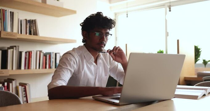 Focused pensive Indian student guy watching online content at laptop, reading electronic article, essay, touching chin, head, thinking, looking at screen, working at research study in library