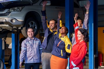 Fototapeta premium mechanics standing with arms raised in celebration and success in a repair shop. Diverse group wearing colorful uniforms, expressing joy and unity in a collaborative automotive work environment.