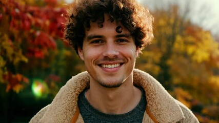 
Young man with curly brown hair smiling brightly, standing outdoors with vibrant autumn leaves in the background