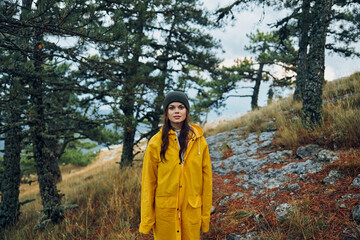 Naklejka premium A young woman in a stylish yellow raincoat standing on a picturesque hill in the midst of a lush forest
