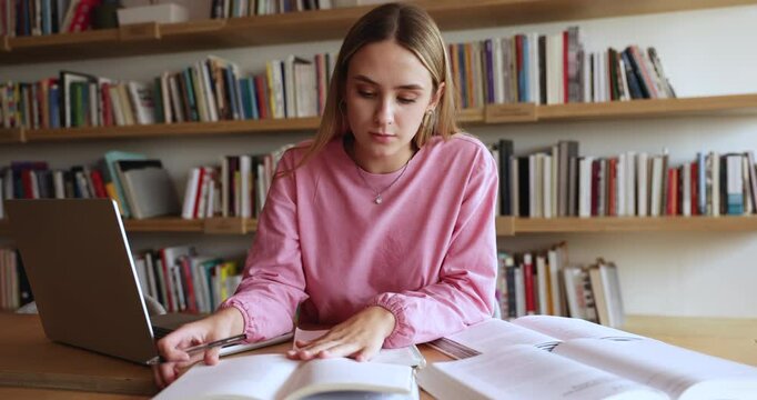 Focused smart Caucasian fresh student girl reading heap of books in library with bookshelves behind, writing notes, making summary for report, research study, sitting at laptop on table