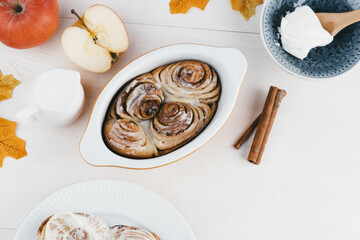 Freshly baked cinnamon rolls sit in a dish beside sliced apples, a glass of milk, and a bowl of ice cream, surrounded by autumn leaves.