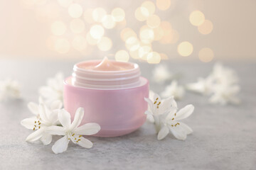 Cream in jar and jasmine flowers on grey table against light background with blurred lights, closeup