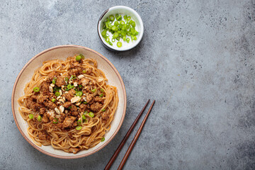 Dan dan noodles are being served with chopsticks and green onions in a bowl, showcasing the...