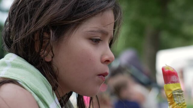 Girl enjoying a popsicle outdoors, wrapped in a towel. The scene reflects the joy of childhood and a refreshing summer treat in a lively park setting