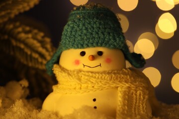 Cute decorative snowman on artificial snow against blurred lights, closeup