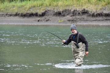 Older, attractive Caucasian man fishing, holding a fishing rod, wearing waders and standing in a river.  Cool, autumn day in salmon season.  Fishing rod is bent with a fish on the hook.