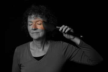 Black and white portrait of a middle aged woman with curly hair. Using a spatula to spread color on her face.