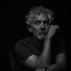 Low key black and white portrait of an attractive older Caucasian man with grey hair and beard. Looking to the right with serious or pensive expression, leaning on his hand