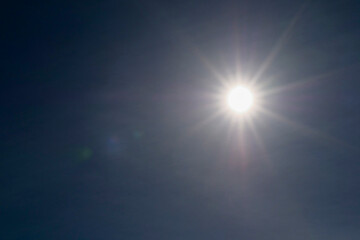 A bright sun is shining through the clouds on a clear day. The sun is the main focus of the image, and the blue sky in the background creates a peaceful and serene atmosphere