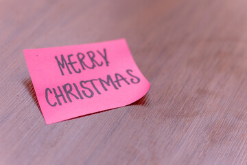 sticky note on a wooden table desk background. written note text is merry christmas