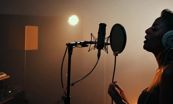 Professional singer singing into microphone close up, recording song in recording sound studio. New hit track release. Woman performing vocal. Live sound. Girl pop performer. Female artist silhouette.