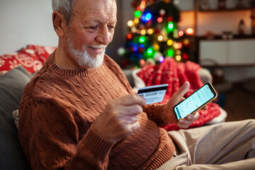 Elderly man using smartphone and credit card for online shopping at home with Christmas lights in background